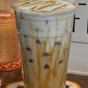 Op de afbeelding: Een hoge, doorzichtige plastic beker gevuld met een gelaagde ijsthee. De drank heeft een cr&egrave;mewitte bovenkant, een karamelkleurige drizzle en een lichtbruine basis met donkerbruine bollen. De beker staat op een decoratieve onderzetter met pompoenthema.