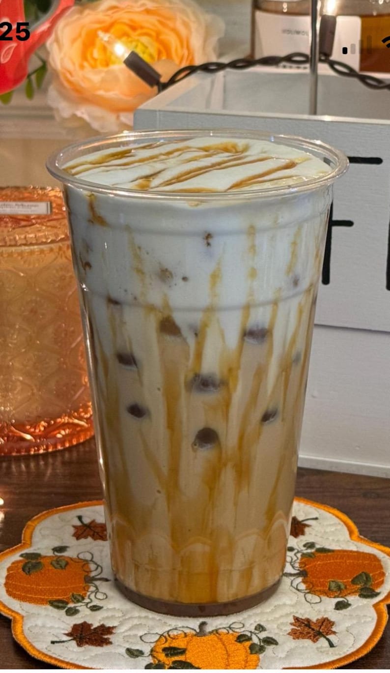 Op de afbeelding: Een hoge, doorzichtige plastic beker gevuld met een gelaagde ijsthee. De drank heeft een cr&egrave;mewitte bovenkant, een karamelkleurige drizzle en een lichtbruine basis met donkerbruine bollen. De beker staat op een decoratieve onderzetter met pompoenthema.