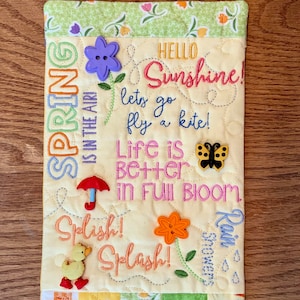 May include: A yellow quilted fabric panel with embroidered text that reads "Spring is in the air! Hello Sunshine! Let's go fly a kite! Life is better in full bloom! Splish! Splash! Rain Showers". The panel is decorated with colorful buttons, including a purple flower, a yellow duck, a red umbrella, a yellow butterfly, and an orange flower.