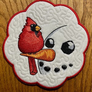 May include: Embroidered patch featuring a red cardinal perched on a snowman's carrot nose. The snowman has black eyes and coal buttons. The patch is white with a red scalloped border and quilted texture.