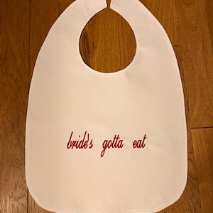 Puede incluir: Babero blanco con cuello curvo y frase bordada en rojo: "bride's gotta eat". El babero est&aacute; hecho de tela suave y est&aacute; dise&ntilde;ado para proteger la ropa durante las comidas. Un divertido accesorio para un evento nupcial.