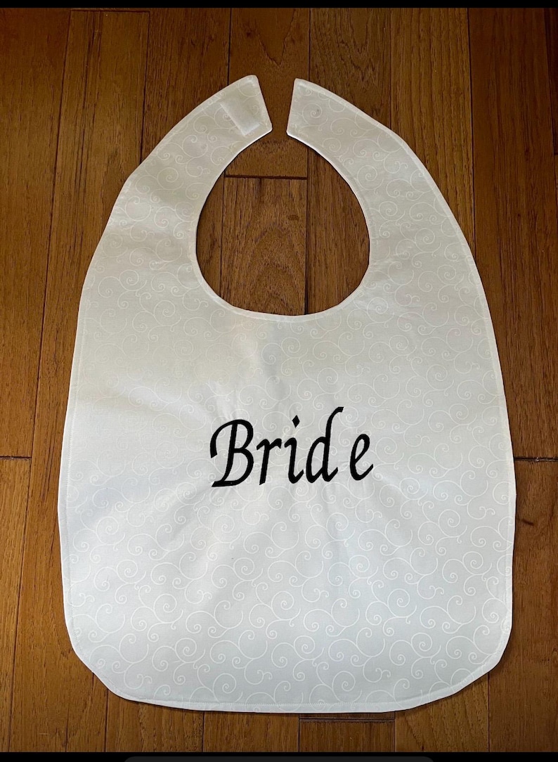 Puede incluir: Babero blanco con un patr&oacute;n de remolinos decorativos y la palabra "Bride" en escritura negra. El babero tiene un escote curvo y una forma rectangular, dise&ntilde;ado para proteger la ropa.