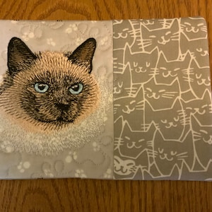 May include: A gray and white fabric pouch with an embroidered image of a Siamese cat on one side and a repeating pattern of gray cats on the other side.