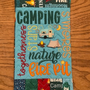 May include: A quilted camping-themed item with the word "CAMPING" embroidered in green. Other embroidered words include "togetherness", "s'mores", "nature", and "fire pit". A small camper, tent, and campfire are also featured.