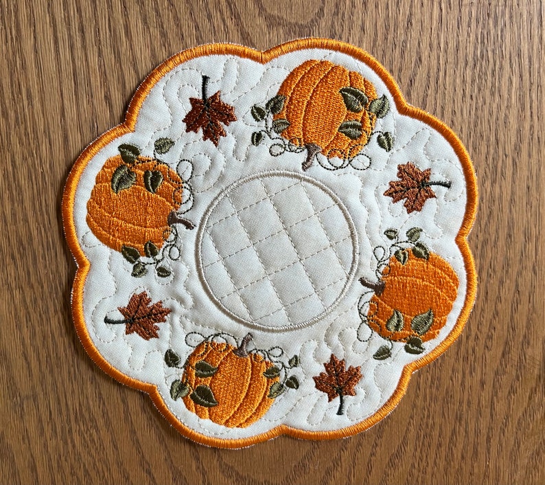 Op de afbeelding: Een cr&egrave;mekleurige, gewatteerde onderzetter met een oranje geschulpte rand. De onderzetter heeft geborduurde oranje pompoenen en bruine bladeren, wat een herfstthema cre&euml;ert. Het midden heeft een gewatteerd cirkelontwerp.