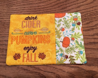 May include: Yellow fabric coaster with embroidered text "drink cider carve pumpkins enjoy fall" and a white fabric panel with a fall-themed print of pumpkins, leaves, and acorns.