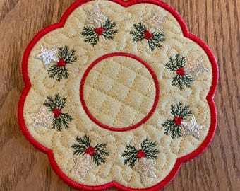 Op de afbeelding: Een ronde, gestikte onderzetter met een rode rand en een ontwerp met een witte ster en groene hulst. De onderzetter is gemaakt van beige stof.