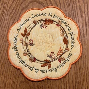 May include: A round, cream-colored fabric coaster with an orange border. The coaster features an embroidered wreath of autumn leaves, acorns, and berries. The text "Autumn leaves & pumpkins please... autumn leaves & pumpkins please" is embroidered around the wreath.