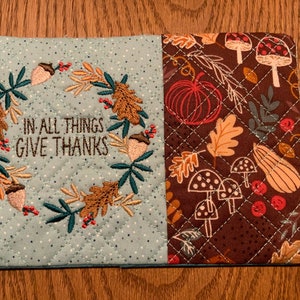 May include: A blue and brown quilted pot holder with an embroidered wreath and the text "In all things give thanks". The brown side features a fall-themed print with pumpkins, mushrooms, and leaves.