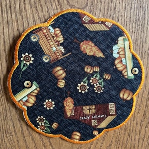 Op de afbeelding: Een decoratieve onderzetter met een geschulpte rand, met een herfstthema. De donkerblauwe stof is bedrukt met pompoenen, zonnebloemen en een wagen. De rand is afgewerkt met een goud-oranje draad. De onderzetter heeft de tekst "Country Acres" erop.