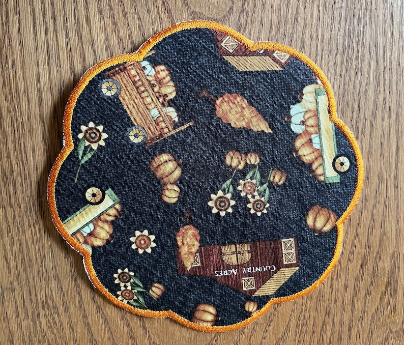 Op de afbeelding: Een decoratieve onderzetter met een geschulpte rand, met een herfstthema. De donkerblauwe stof is bedrukt met pompoenen, zonnebloemen en een wagen. De rand is afgewerkt met een goud-oranje draad. De onderzetter heeft de tekst "Country Acres" erop.