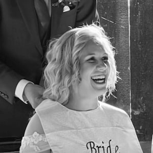 Puede incluir: Foto en blanco y negro de una persona sonriente que lleva un babero blanco con la palabra "Bride" bordada. La persona tiene el pelo rubio rizado y lleva un vestido con detalles de encaje. Una persona con traje est&aacute; detr&aacute;s de la persona.