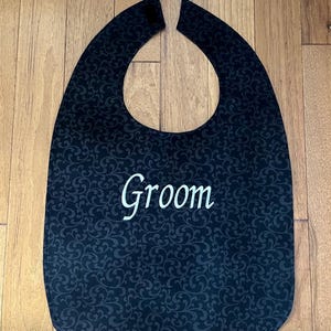 Puede incluir: Un babero negro con un patr&oacute;n de desplazamiento decorativo y la palabra "Groom" bordada en blanco. El babero tiene forma curva y cierre de velcro en el cuello.