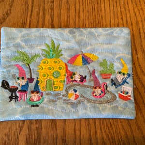 May include: Embroidered fabric pouch with a blue background and a scene of gnomes enjoying a summer day at the beach. The gnomes are wearing sunglasses and are relaxing in a pool, grilling, and floating on a flamingo.