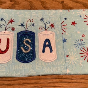 Puede incluir: Una bolsa de tela azul con fondo blanco y fuegos artificiales bordados en rojo, blanco y azul y las letras "USA" en el centro.