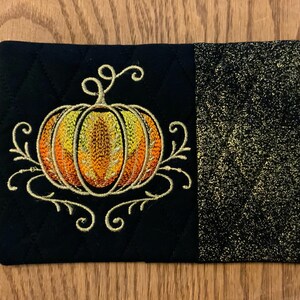 May include: A black quilted fabric coaster with a gold embroidered pumpkin design. The coaster has a textured black fabric on the back.