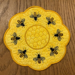 May include: A yellow flower-shaped coaster with a honeycomb design in the center and eight embroidered black and yellow bees around the edge.