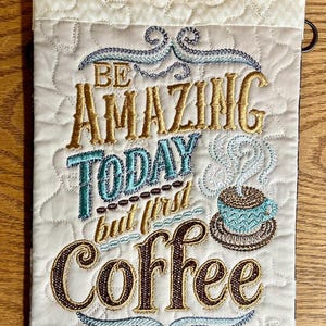 May include: Embroidered wall hanging with the text "Be Amazing Today but first Coffee". The design features a coffee cup, steam, and decorative flourishes. The background is quilted with a cream-colored fabric.