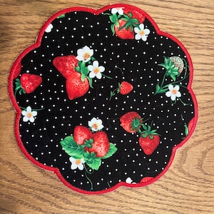May include: A scalloped fabric coaster featuring a black background with a white polka dot pattern, red strawberries, green leaves, and small white flowers. The edge of the coaster is finished with a vibrant red stitched border.