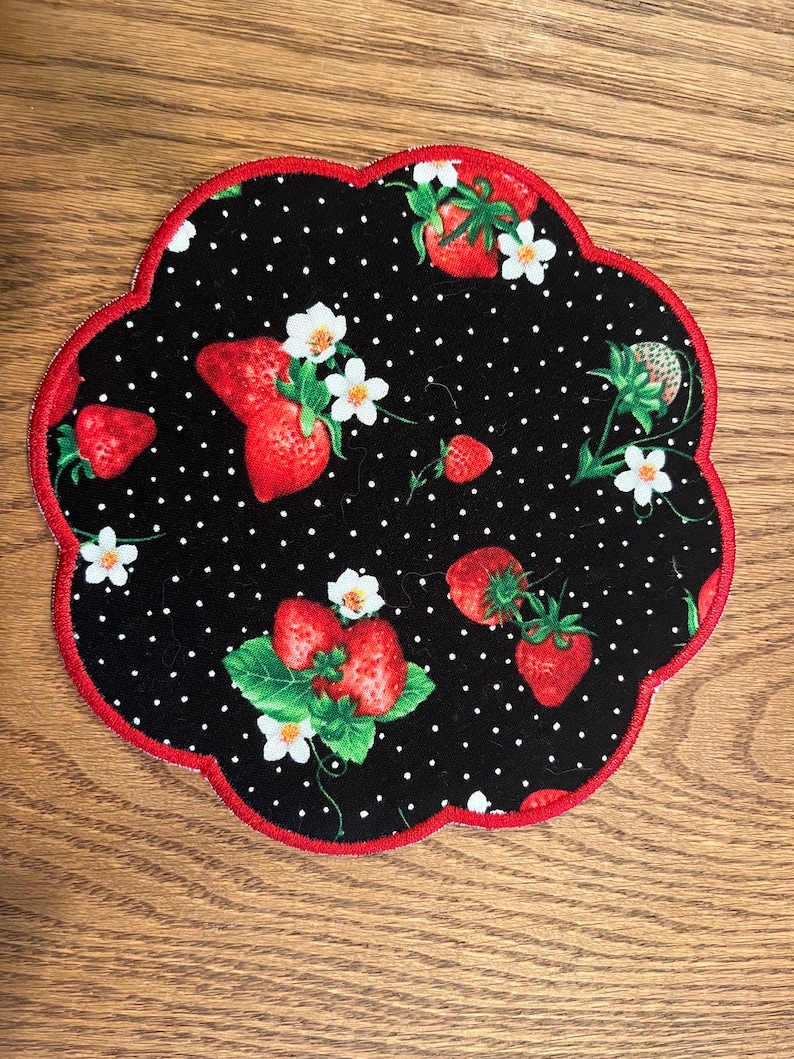 May include: A scalloped fabric coaster featuring a black background with a white polka dot pattern, red strawberries, green leaves, and small white flowers. The edge of the coaster is finished with a vibrant red stitched border.