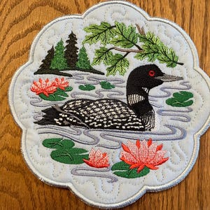 May include: A blue and white quilted coaster with an embroidered loon swimming in a pond with lily pads and green trees.