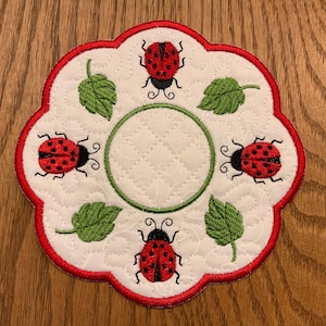 May include: Embroidered white coaster with a red scalloped border. The coaster features a quilted pattern, a green circle in the center, and is decorated with red ladybugs and green leaves. A decorative item for the home.