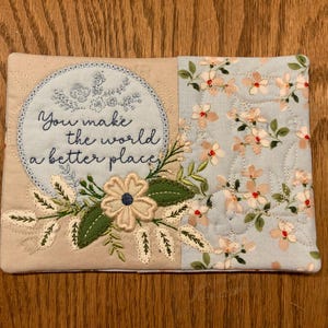 May include: A beige and blue quilted coaster with a floral design and the text "You make the world a better place."