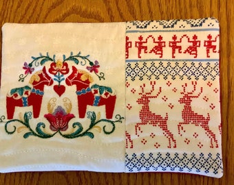 May include: A white fabric coaster with a red and green embroidered design of two Dala horses and a flower. The other side of the coaster features a white fabric with a red and blue embroidered design of two reindeer.