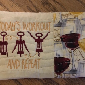 May include: A white fabric coaster with the text "Today's Workout and Repeat" embroidered in gold thread. Three red wine bottle openers are embroidered on the left side of the coaster. The right side of the coaster features a white fabric with a pattern of wine glasses and grapes.