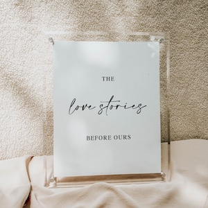 May include: A clear acrylic sign holder with a white card featuring the text "THE love stories BEFORE OURS" in black script. The sign is displayed on a beige fabric surface, suitable for weddings or special occasions.