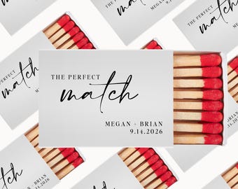 Könnte beinhalten: Weiße Streichholzschachteln mit roten und weißen Streichhölzern. Die Schachteln tragen den Text "The Perfect Match" in handschriftlicher Schrift und die Namen Megan und Brian mit ihrem Hochzeitsdatum.