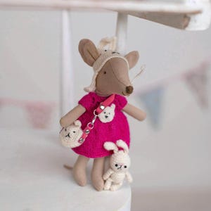 May include: A handmade plush mouse doll wearing a bright pink dress, a cream-colored hat, and carrying a small purse. A small white crocheted bunny doll stands beside the mouse. The mouse doll has a pink strap with a bear-shaped detail.