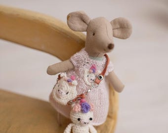Op de afbeelding: Een handgemaakte stoffen muispop met een roze jurk, een bijpassende handtas en een kleine witte pop. De muis heeft grote oren en een bruine neus. De handtas en de kleine pop hebben een bloemenpatroon.