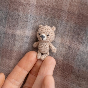 May include: A small, crocheted teddy bear in a light brown color. The bear has a dark gray nose and black eyes. The bear is approximately 2 inches tall and is held in a person's hand. The background is a soft, patterned fabric.
