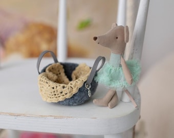 May include: A small gray felt basket with a crocheted trim sits on a white wooden chair. A gray and brown mouse doll wearing a light green tulle skirt is sitting on the chair.