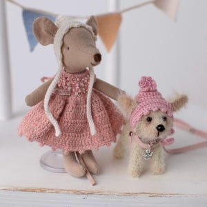 Può includere: Una piccola bambola di topo con un vestito rosa all'uncinetto con bordi bianchi e un cappello bianco all'uncinetto. Una piccola bambola di cane bianco all'uncinetto con un cappello rosa all'uncinetto e un guinzaglio è in piedi accanto alla bambola di topo.