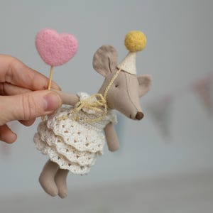 May include: A handmade mouse doll in a white crocheted dress and a party hat with a yellow pom-pom. The mouse holds a pink felt heart on a stick. The doll is a whimsical, handcrafted item.