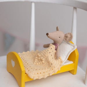 Puede incluir: Un pequeño juguete de ratón beige en una cama de madera amarilla con una almohada blanca y una manta de ganchillo. La cama tiene un recorte en forma de corazón. La manta es de color amarillo claro. Un artículo decorativo encantador.