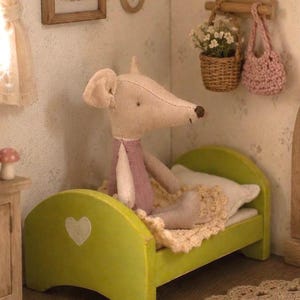 May include: A charming doll's house interior with a stuffed mouse in a lime green bed. The bed has a heart detail. A framed picture of a mouse is on the wall. A small basket of flowers and a pink crocheted bag are also present.