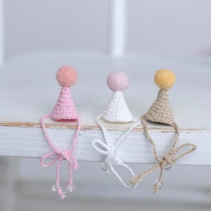 Op de afbeelding: Drie kegelvormige miniatuurhoedjes van gehaakt garen, elk bekroond met een klein vilten pomponnetje. De hoedjes zijn roze, wit en beige en hebben dunne, bijpassende kinbandjes met kleine doorzichtige kraaltjes aan de uiteinden van de koordjes.