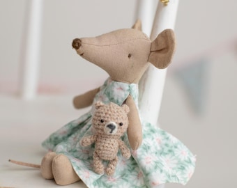 May include: A handmade stuffed mouse doll wearing a floral dress, with a small crocheted teddy bear. The mouse is light brown with large ears and a long tail. The dress is light green with white flowers. The toys are sitting on a white wooden chair.