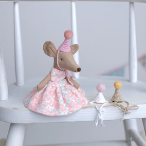 May include: A fabric mouse doll dressed in a pink floral dress and a pink party hat. Two miniature party hats, one white and one beige, are also visible. The doll is on a white wooden chair.
