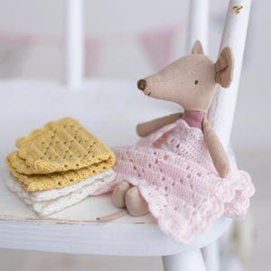 May include: A soft toy mouse in a pink crocheted dress sits on a white chair. Next to the mouse are stacked yellow and white crocheted coasters. The mouse has tan fabric ears and a long nose.