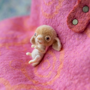 May include: A handmade, miniature felted creature with a light brown head, large ears, and a cream-colored body. It is lying on a textured pink surface. The creature has large, dark eyes and is posed with its arms and legs extended.