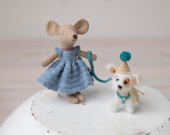 May include: A handmade mouse doll wearing a blue dress and holding a teal leash, walking a small white dog with a party hat. The dog has brown spots and a teal collar. Both are on a white surface.