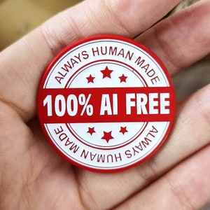 Op de afbeelding: Een ronde rode sticker met witte tekst die "ALWAYS HUMAN MADE 100% AI FREE ALWAYS HUMAN MADE" luidt, met vijf witte sterren rond de tekst.