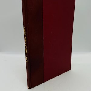 May include: A hardback book with a burgundy cover and spine. The spine displays the title "PEKOO, THE CAT WHO TALKS" in gold lettering. The book is closed and upright against a white backdrop.