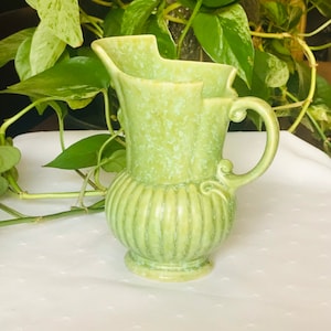 May include: A green ceramic jug with a speckled glaze and a fluted design. The jug has a handle and a wide mouth.