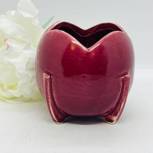 May include: A small, heart-shaped, burgundy ceramic planter with three small feet. The planter has a glossy finish and a wide opening. A white flower is partially visible in the background.