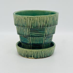 May include: A green ceramic planter with a matching saucer. The planter has a textured, basket-weave design. The planter is approximately 15 cm in diameter and 13 cm tall. The saucer is about 15 cm in diameter.
